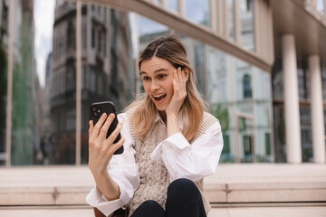 Fototapeta premium Beautiful smiling fashionable businesswoman sitting on the stairs in front of business center and make selfie, hold mobile phone, look happy. Amazed woman wear beige knit vest, white shirt and bag.