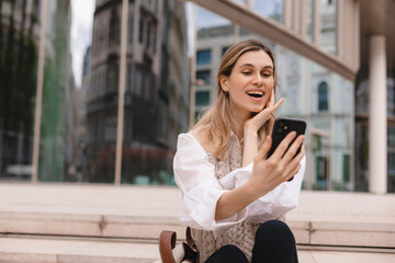 Fototapeta premium Beautiful smiling fashionable businesswoman sitting on the stairs in front of business center and make selfie, hold mobile phone, look happy. Amazed woman wear beige knit vest, white shirt and bag.
