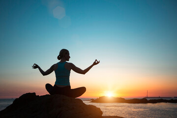 A silhouette of a middle-aged woman in a lotus pose doing yoga on the ocean at dusk.