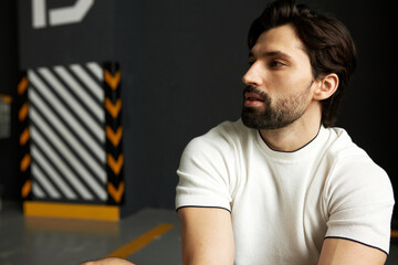 Closeup indoor portrait of attractive fitness instructor having rest after training session in white t-shirt on his muscled and toned body and arms, looking to the right at copy space background