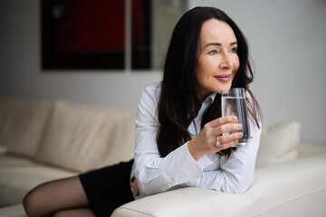 Attraktive Frau im Best-Ager Alter mit einem Glas Wasser zuhause auf der Couch 