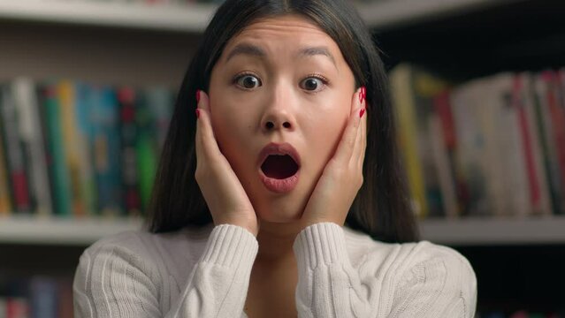 Close-up surprised young asian woman in shock with frightened expression opens mouth shocked by bad news feeling stress female student teacher standing in library near bookshelf worries about problem