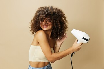 Woman dries curly afro hair with blow dryer, home beauty care styling products hair, smile on beige...