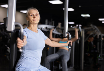 Fototapeta na wymiar Concentrated motivated sporty elderly woman exercising chest muscles on butterfly machine during upper body strength training in gym