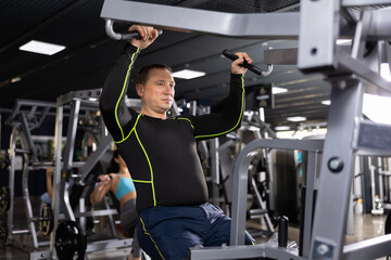 Fototapeta na wymiar Concentrated adult man in sportswear working out in modern gym, performing exercises for back muscles building on lat pull down lever machine. Fitness and bodybuilding concept..