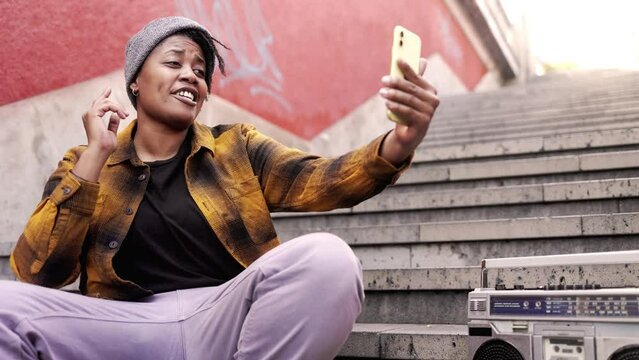 Influencer African woman streaming live on social media sitting on stairs in urban street outdoors