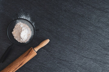 A sieve with flour and a rolling pin on a black stone table.