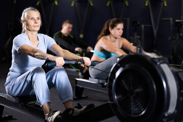 Concentrated sporty senior woman working out on rowing machine during total-body workout in gym....