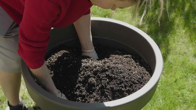 mixing soil for planting flowers and green plants in big grey flower pot outdoors spring sunny day. gardening woman hands in gloves mix stir soil soft ground prepare planting replanting plants