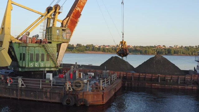The crane loads the sand onto the ship. The barge ship floats on the water, coastline in the background. Drone flying over barges