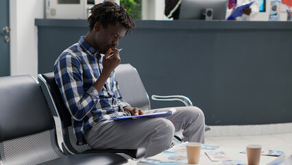 Sick young adult filling in checkup report files on clipboard, writing registration form before starting appointment with physician. Male person using medical papers in waiting room.