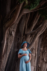 Fototapeta na wymiar Portrait of middle-aged pregnant woman in denim blue dress, hat. The future mother stands among the huge, brown trees. Travel during pregnancy. Maternity and woman pregnancy concept.