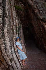 Pregnant millennial woman in blue dress stands among the rocks on the seashore, hugging belly with her hands. Girl in hat on background of textured wall. Natural beauty concept. Copy space, traveller