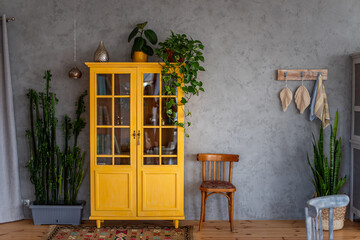 Deep yellow wooden bookcase against gray concrete wall. Around green pots with home flowers. Scandinavian loft style apartment rustic design. Macrame elements. Trendy, botanical room decor. Copy space © farmuty
