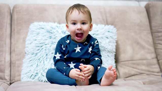 Beautiful baby boy sits on the sofa and yawns. Little child watching TV attentively in the living room.