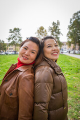 Two smiling friendly Asian women pose back to back in a public park