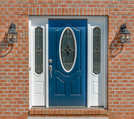 Close up view of elegant blue wooden door with oval stained window, with white sidelights, silver brass color door knob, iron wall light on red brick facade