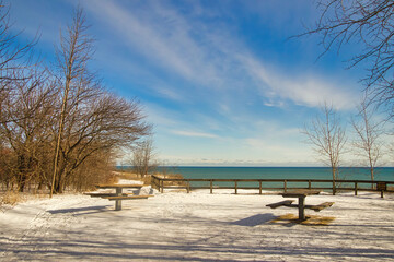 Fototapeta na wymiar Beneath a mostly blue Winter sky, two empty picnic tables along a hiking trail on a snow-covered bluff offer a view of Lake Michigan at Lion's Den Gorge, near Grafton, WI.