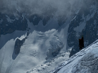 A lone snowboarder ascends a steep, icy ridge under dramatic, shadowy peaks. The harsh alpine...