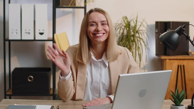 Young business woman working on laptop computer smiling friendly at camera and waving hands gesturing hello, hi, greeting or goodbye, welcoming with hospitable expression at home office workplace desk