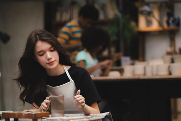 Portrait of young Asian girl wearing apron preparing pots and sculptures using mud and and clay and showing thumbs up in a workshop studio while smiling and looking at camera
