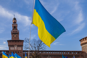 Flag with yellow and blue striped colors of Ukraine waving in the wind with a blue sky and sun. In the background the Castello Sforzesco in Milan.