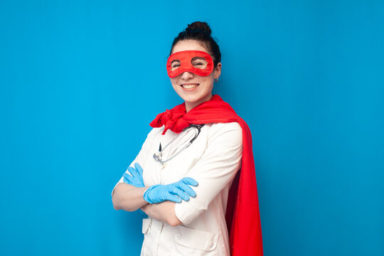 cheerful young girl doctor in uniform in superman costume on blue background, female nurse in superhero mask