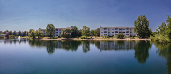 Panorama of apartments reflected on a small lake in Boise, Idaho. Small lake at the front with reflections of apartments, trees, and clear indigo sky above.