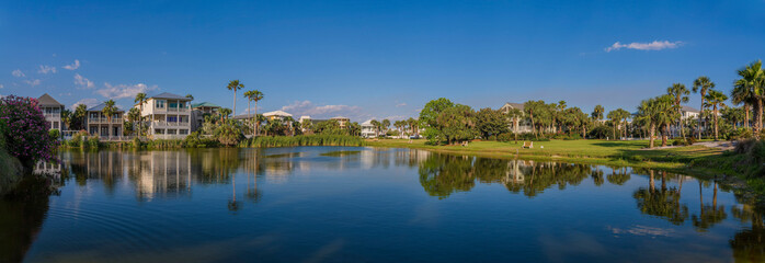 Destin, Florida- Panorama of a lake in a residential area with reflections of blue sky. Lake with field and grasses on the shoreat the front of the three-storey houses. - Powered by Adobe