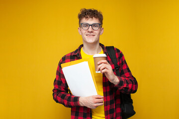 curly guy student in glasses with a backpack and papers holds coffee on a yellow background and...