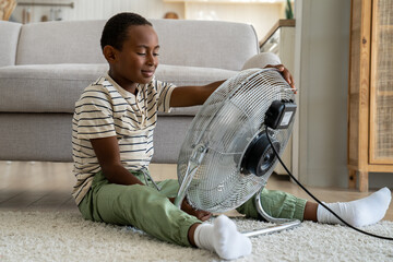 Happy African boy refreshing with big fan sitting on floor in living room alone. Black preschool...