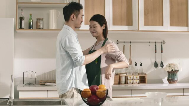 loving caring asian husband helping wife put on apron in kitchen at home