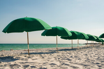Green beach umbrellas in a row at the beach in Destin, Florida. Umbrellas on the white sand near the ocean water against the clear blue skies at the background.