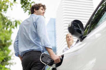 Focus hand insert EV charger to electric vehicle at public charging point in car park with blur business people in backdrop, eco-friendly lifestyle by rechargeable car for progressive concept.