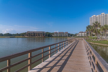 Fototapeta na wymiar Perspective view of footbridge over Stewart Lake heading to hotels and apartments in Destin, Florida. Lakefront apartments and hotels with low-rise front and high-rise back structures against sky.