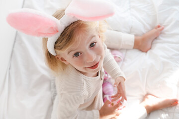 Happy caucasian girl two years old wearing rabbit bunny ears playing with eggs sitting on bed...