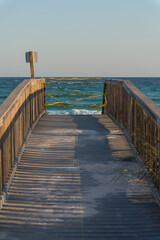 Vertical shot view of a wooden boardwalk with yellow caution tape in Destin, Florida. Walkway with wooden railings and sign post on the left and a view of blue ocean and sky background.