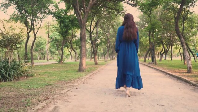 Desi woman is slowly walking in a park in a blue dress and heels, leisure and relaxing activities concept.