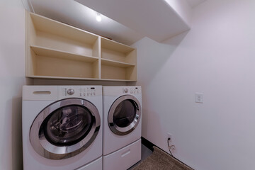 Small laundry room with front load washing machines under the wall shelves. Laundry room interior with white walls and carpet flooring.