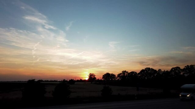 Wind turbines against orange horizon at sunset; long shot from driving car