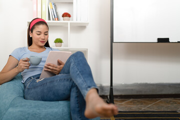 Fototapeta premium Young woman, sitting on a pouffe, using a digital tablet and drinking a cup of coffee.