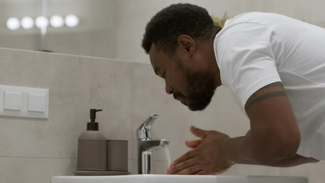 Personal hygiene habit. Young sleepy african american guy washing his face with water in morning, waking up at bathroom