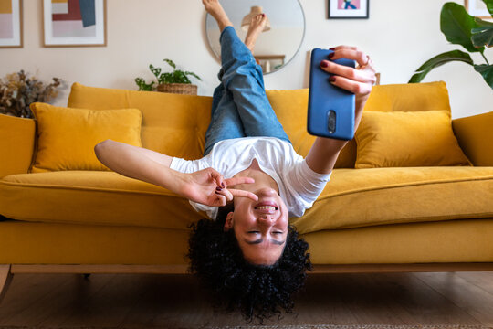 Joyful African American woman lying upside down on the sofa takes selfie with smart phone. Social media.