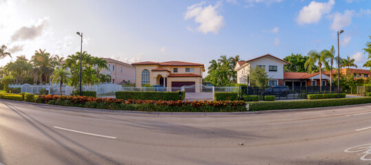 Miami, Florida- Panorama of gated mediterranean houses against the sunny sky. There is a road at the front with bushes and street lights at the front of the houses with gates and fences. - obrazy, fototapety, plakaty