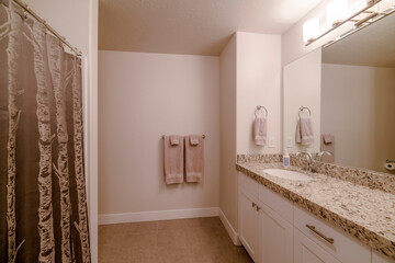 Interior of residential bathroom with view of sink above counter top and cabinet. A shiny shower curtain, bright wall lights, and towels hanging on towel ring and rack can be seen inside the restroom.