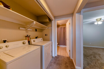 Laundry room with wall shelves beside the bathroom near the empty room. There is a carpeted empty room on the right and laundry units at the entrance of the tiled bathroom on the left.