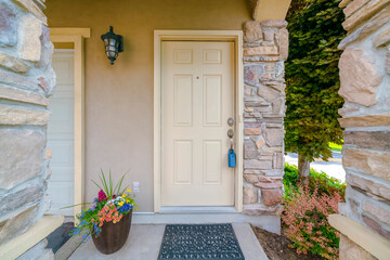 House entrance with cream front door with lockbox near the plants and tree on the side. There are flowers on a pot on the left near the garage door and front door on the right.