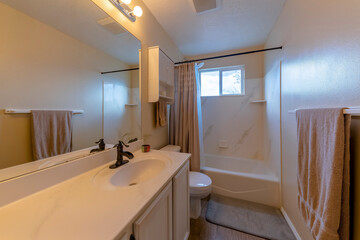 Fototapeta na wymiar Bathroom with a vanity sink and hanging light brown shower curtain and towels. There is a sink with antique faucet near the toilet with wall cabinet beside the tub shower with window at the back.