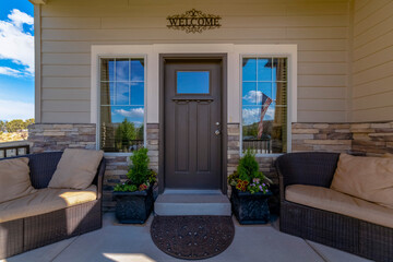 Front view of a home with glass paned gray front door and relaxing porch sofa. Reflection of an American flag and vibrant blue sky on this sunny day can be seen on the window.
