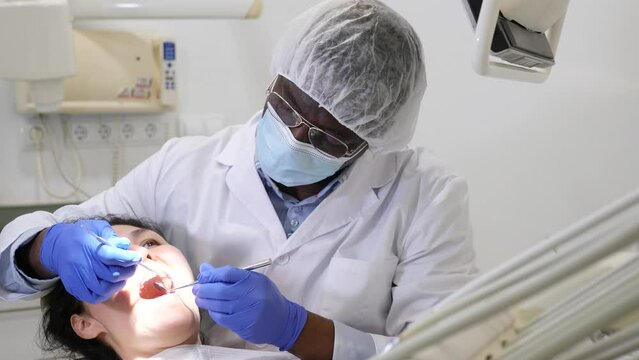 African-american man orthodontist using tools and doing oral care inspection on Asian woman patient to cure toothache. 
